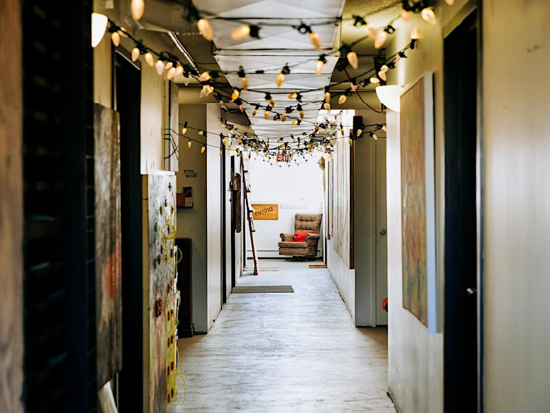 Hallway with stringlights hung on the ceiling with doors on either side and leading to room with a leather armchair.
