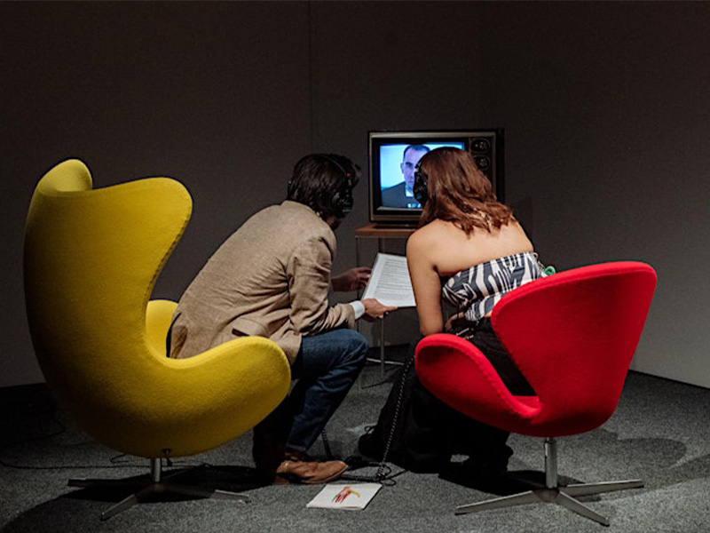 A man sitting in a yellow curved chair, a woman next to him in a red curved chair, both leaning forward towards a tv screen in the background