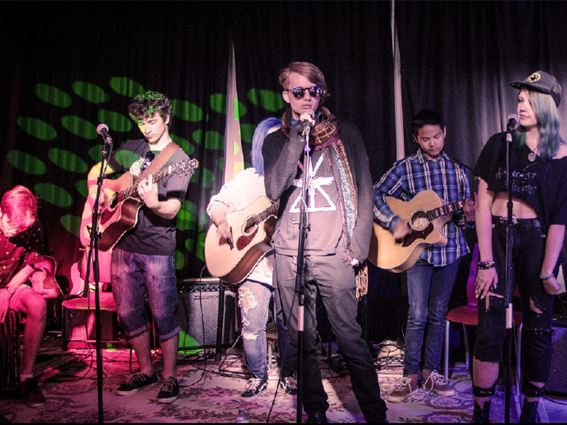 Six youths on stage performing in front of microphones with instruments