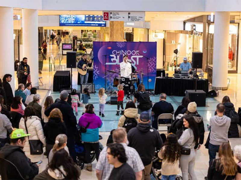 A crowd of people gather around a performer standing on a small stage with a microphone. The purple signage behind him reads Chinook Blast
