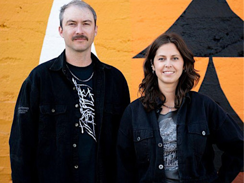 A man and a woman standing side by side, each wearing a dark jacket over a tshirt, Behind them an orange background