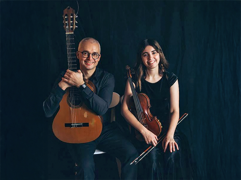 A man holding a guitar upright on his lap smiles while sitting next to a woman with a violin under her arm and holding a bow. They are wearing dark clothing against a dark background.