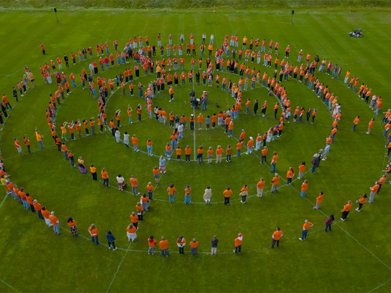 A group of many people stand on a green field forming the shape of a medicine wheel. The people are primarily wearing orange clothing.