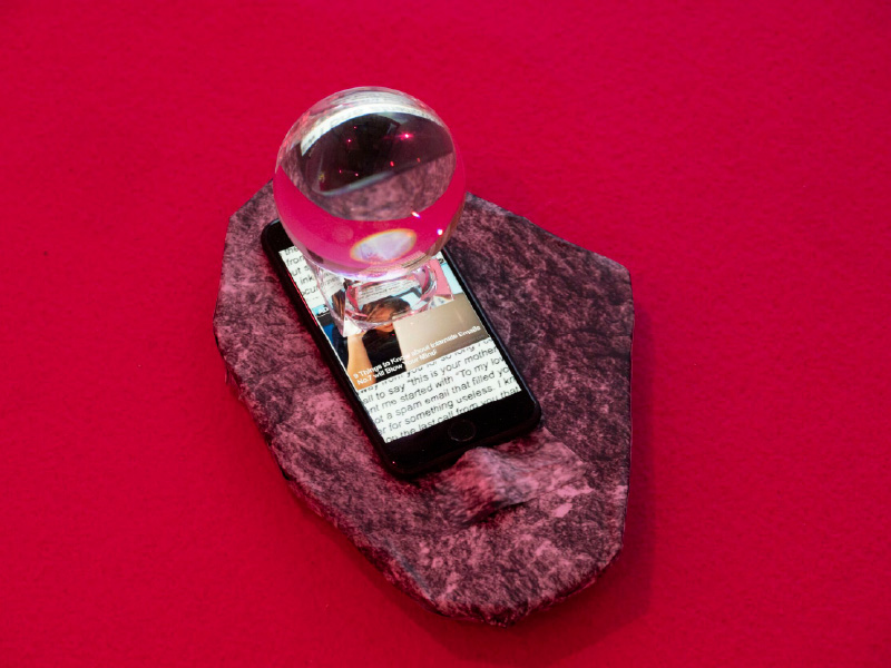 Smartphone sits between a flat piece rock and small glass orb on top. All against on red-coloured backdrop