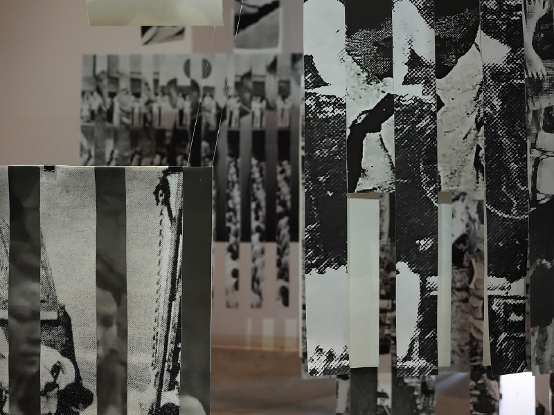 Large black and white images suspended from ceiling at varying depths and heights, At closer look, it appears that images are made up of multiple vertical strips of black and white images stitched together